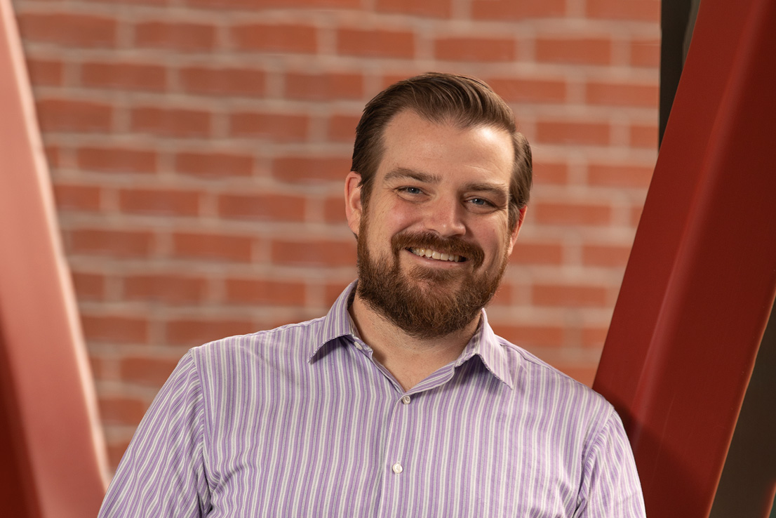 A headshot of Jon Mathis with a full beard wearing a light purple striped dress shirt and smiling warmly while standing against a brick wall with red metal beams